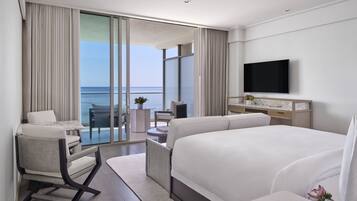 A modern hotel room with a large bed, a TV, a balcony with a view of the sea, and a seating area with a table and chairs.