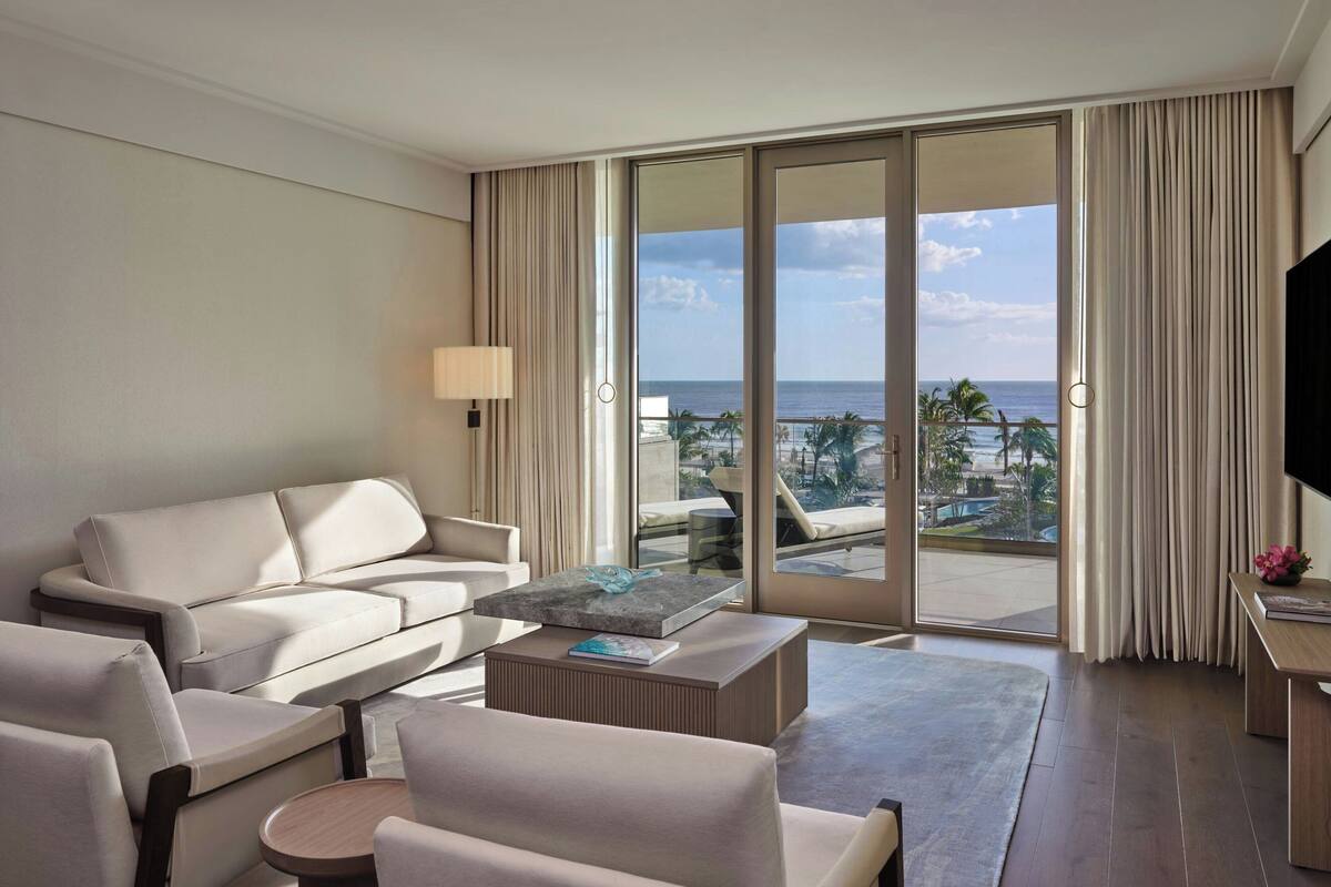 Ein modernes Hotelzimmer mit einer Couch, Sesseln, einem Couchtisch und einem Fernseher. Das Zimmer verfügt über eine große Schiebetür, die auf einen Balkon mit Meerblick führt.