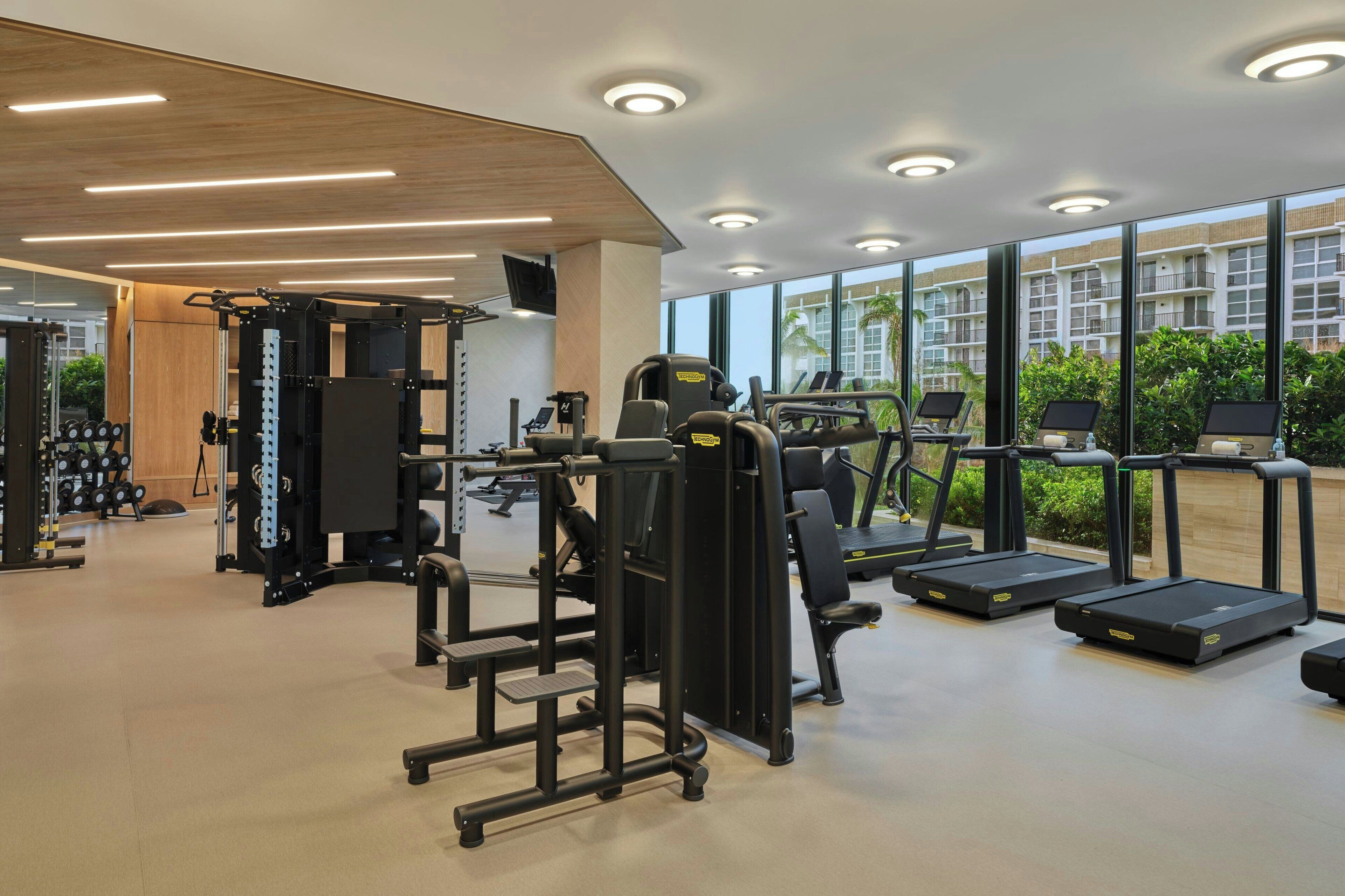 A modern gym with various exercise equipment, large windows, and a view of a residential area.