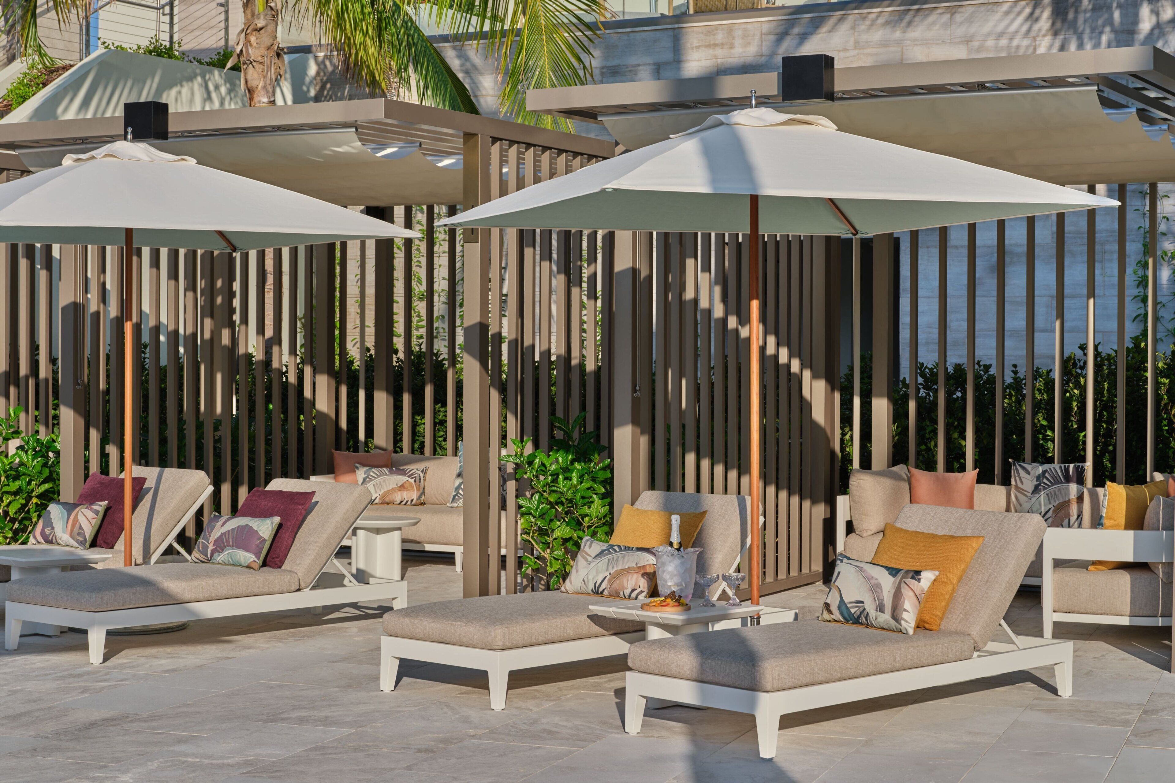 A sunlit outdoor lounge area with white lounge chairs and umbrellas, surrounded by vertical wooden slats and greenery.