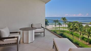 A balcony with a sofa, a table, and a bench, overlooking a beach with palm trees and a clear sky.