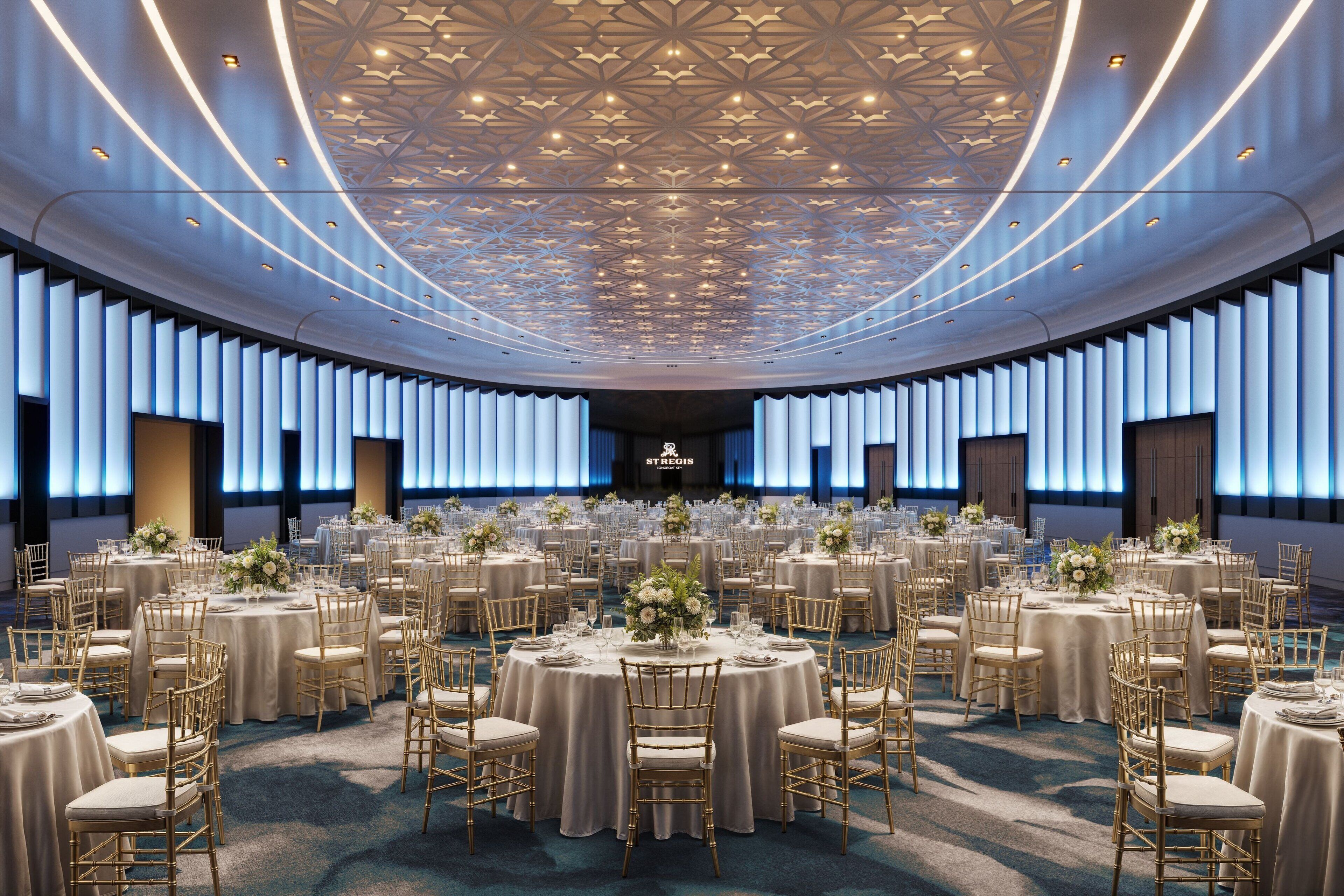 A large, circular banquet hall with round tables and gold chairs, featuring a modern ceiling design and a stage at the front.