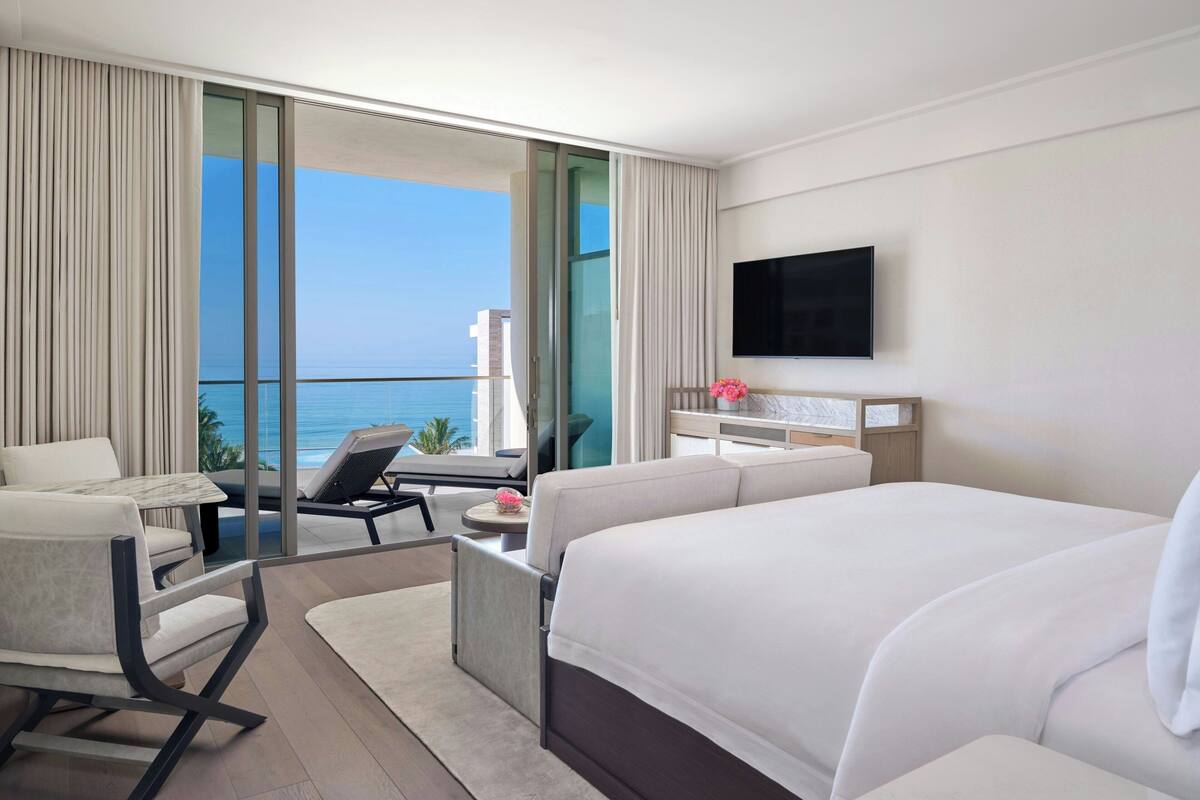 Ein modernes Hotelzimmer mit einem großen Bett, einem Schreibtisch, einem Sessel und einem Balkon mit Meerblick.