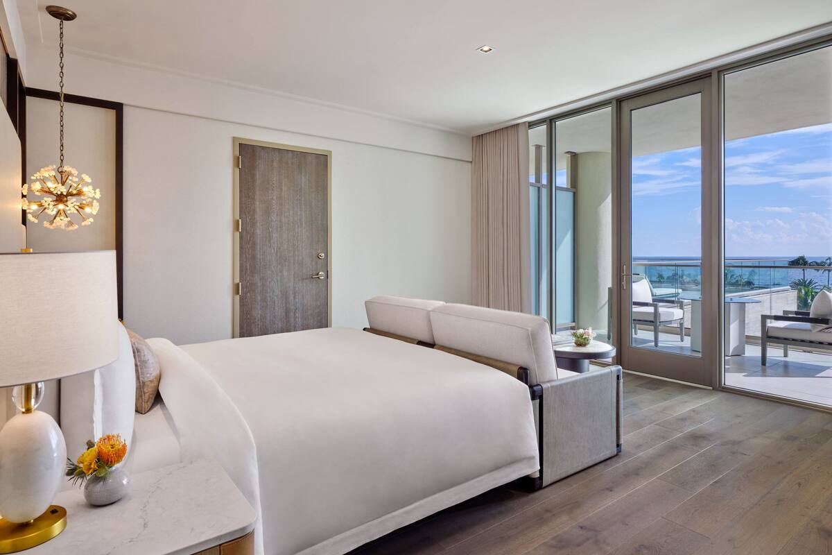 Ein modernes Hotelzimmer mit einem großen Bett, einem Nachttisch mit Lampe, Blick auf einen Balkon mit Sitzgelegenheit und einer großen Schiebeglasdoor.