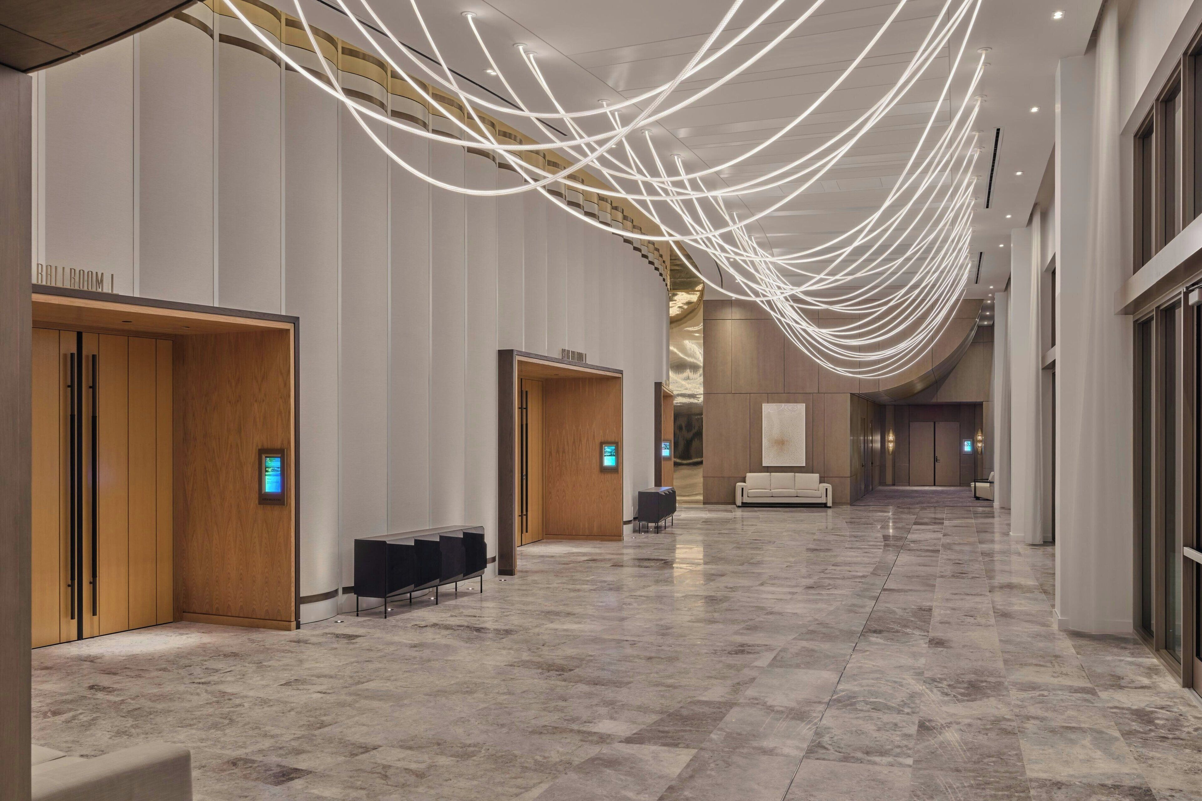 Een ruime hotel lobby met een marmeren vloer, houten deuren en een moderne lichtinstallatie.