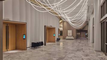 Een ruime hotel lobby met een marmeren vloer, houten deuren en een moderne lichtinstallatie.