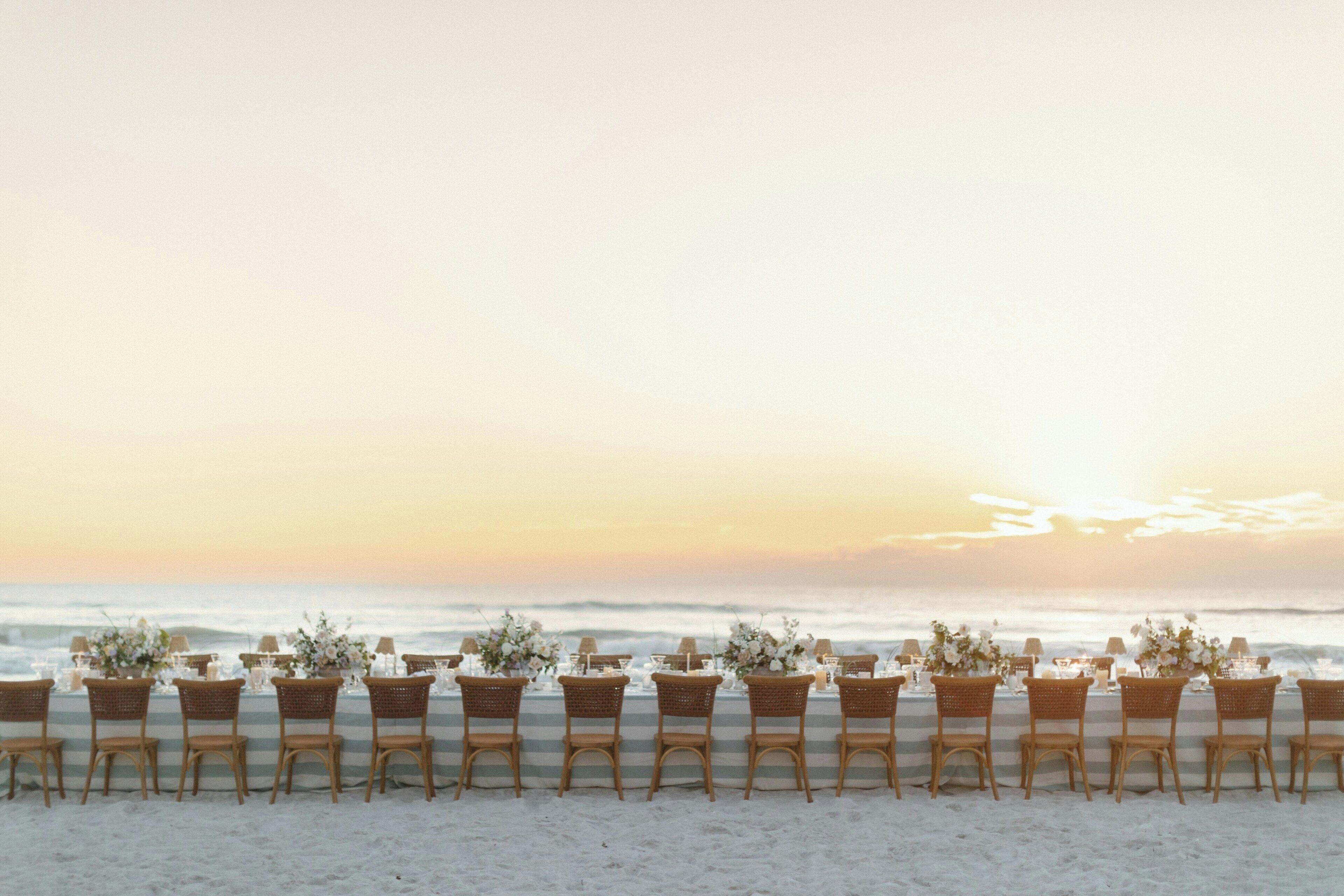 A beachside dining setup with wicker chairs and tables, each adorned with floral centerpieces, overlooking the ocean.