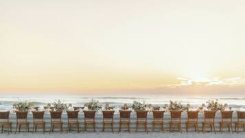 A beachside dining setup with wicker chairs and tables, each adorned with floral centerpieces, overlooking the ocean.