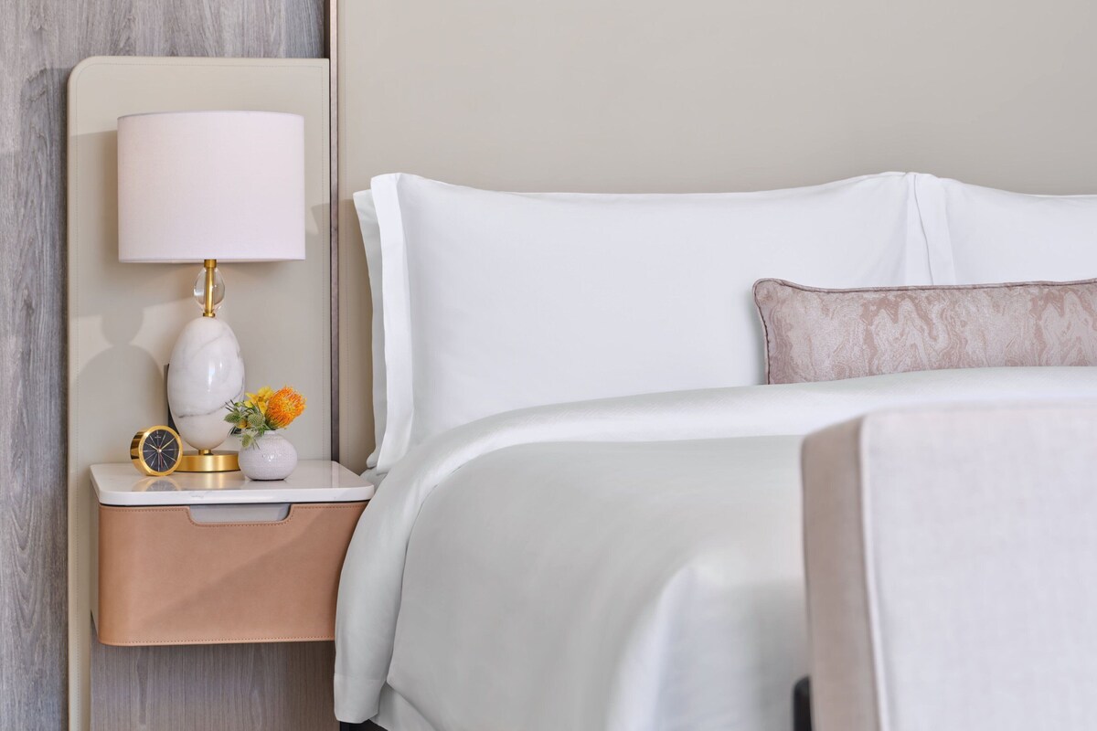 A neatly made bed with white linens and a single decorative pillow, a bedside table with a lamp, and a small vase with flowers.