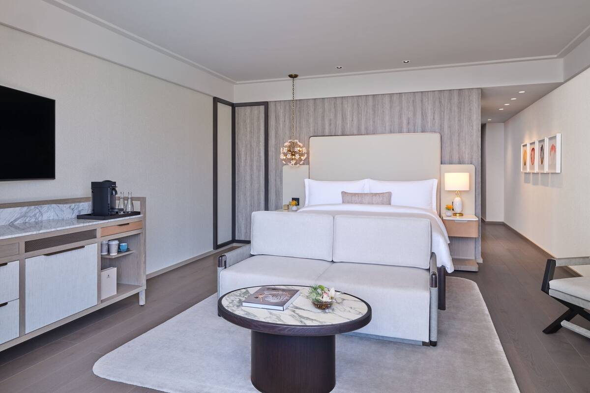 Ein modernes Hotelzimmer mit einem großen Bett, einem Nachttisch mit Lampe, einem runden Couchtisch und einem an der Wand befestigten Fernseher.