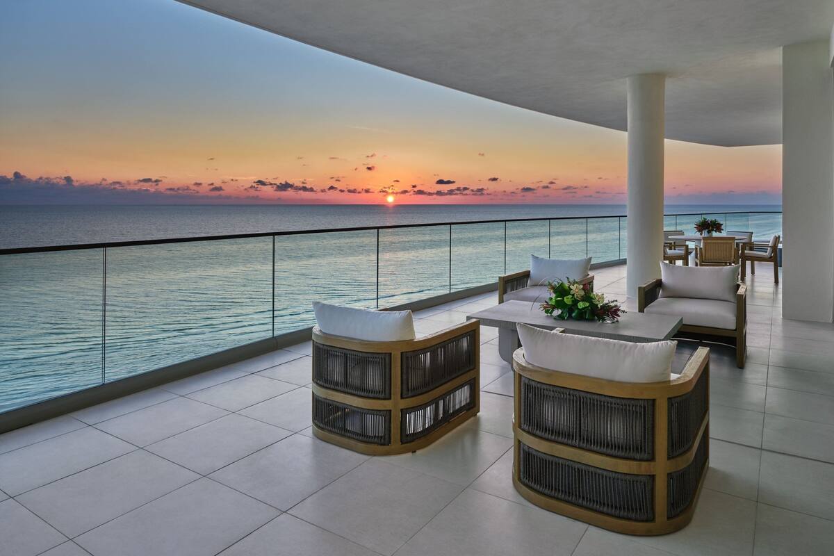 Ein Balkon mit Korbmöbeln, einem Tisch mit einer Vase und Blick auf den Sonnenuntergang über dem Meer.