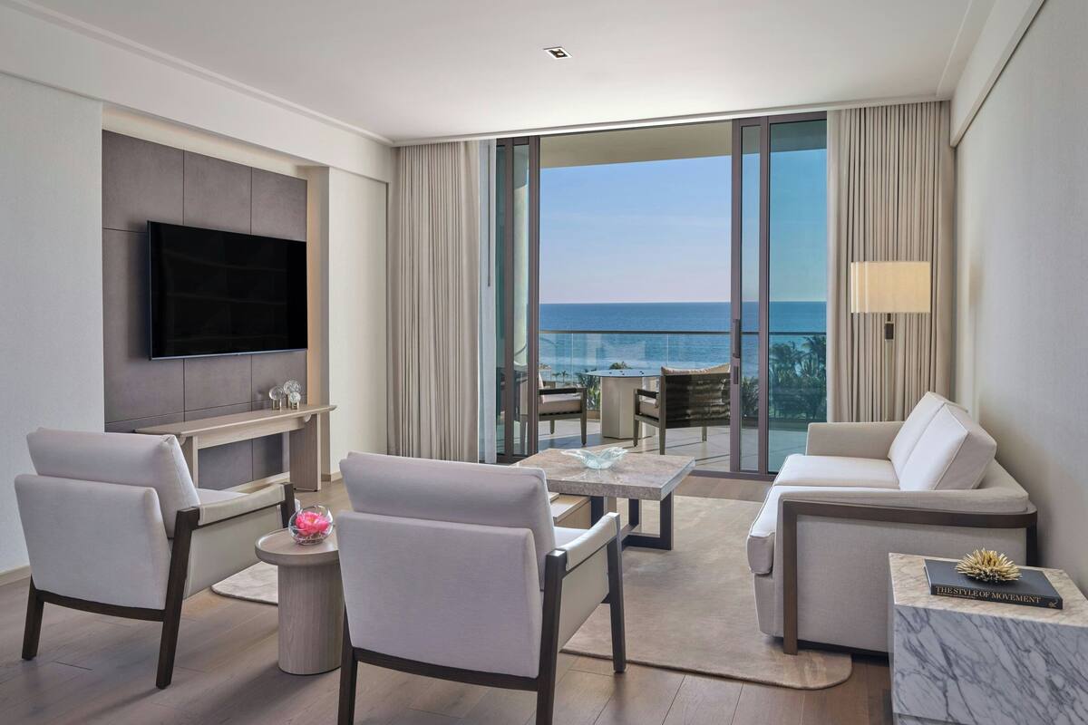 Ein modernes Hotelzimmer mit Flachbildfernseher, einer Couch, zwei Sesseln, einem kleinen Couchtisch und einem Balkon mit Meerblick.