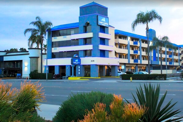 A multi-story hotel with a blue and white facade, palm trees, and a parking lot. - Capri Suites Anaheim (Anaheim)