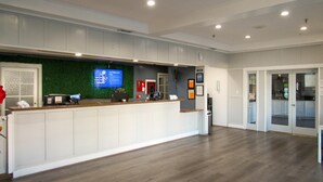 A reception area with a wooden counter, a green wall, and a large screen displaying information. - Capri Suites Anaheim (Anaheim)