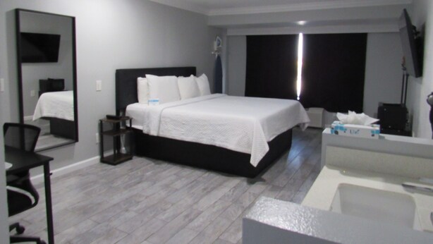 A modern hotel room with a large bed, a desk, a chair, a mirror, and a bathroom with a bathtub. - Capri Suites Anaheim (Anaheim)