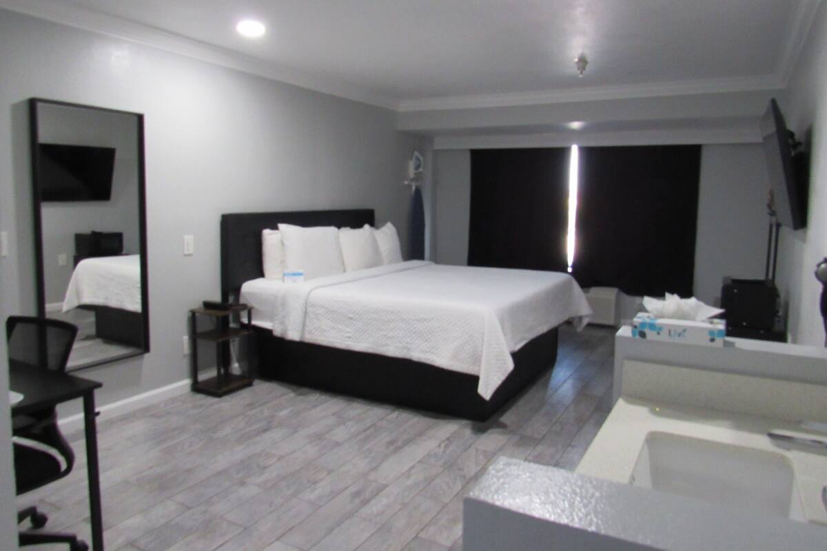 A modern hotel room with a large bed, a desk, a chair, a mirror, and a bathroom with a bathtub.