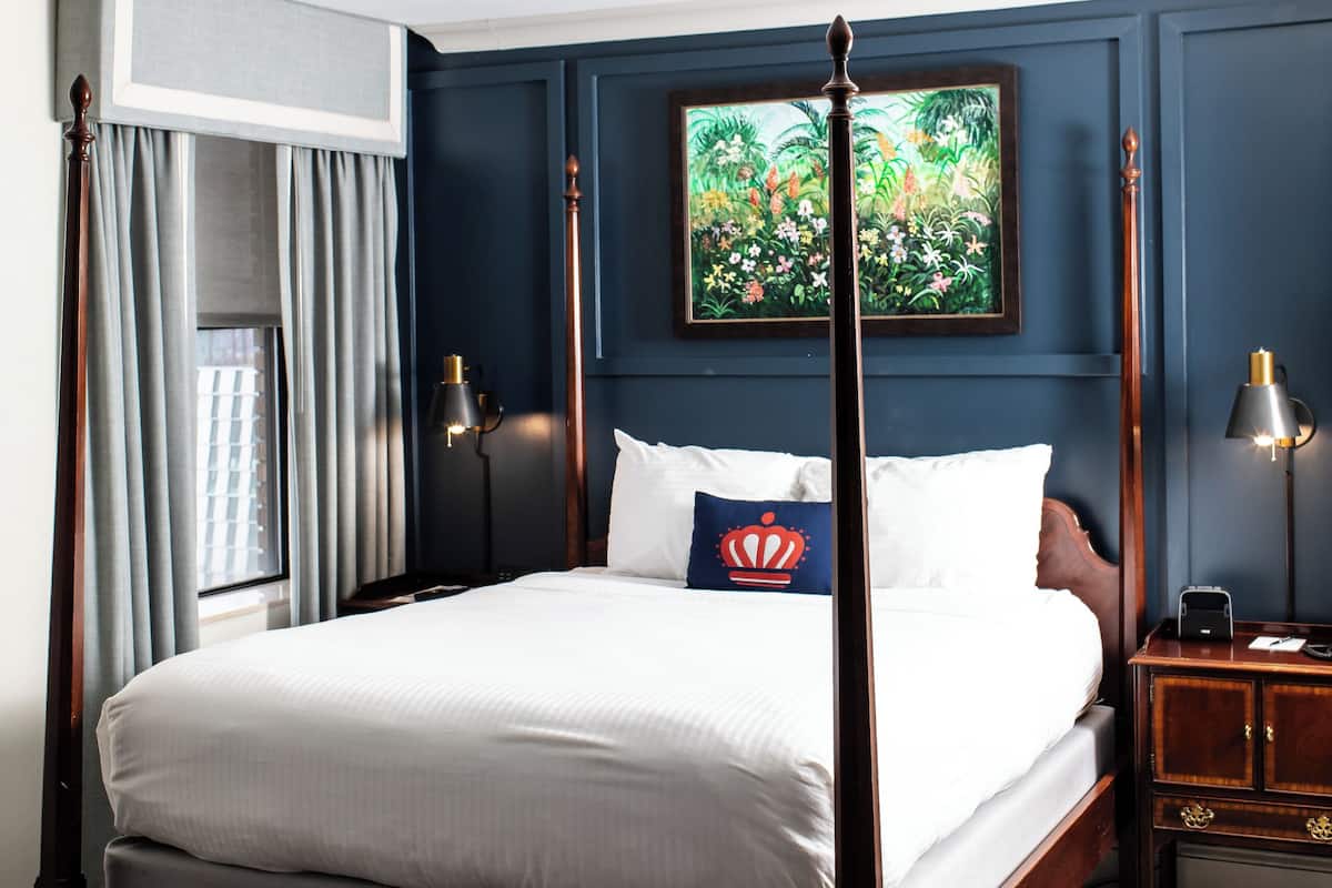 A four-poster bed with a headboard, a nightstand with a lamp, a painting on the wall, and a window with curtains.