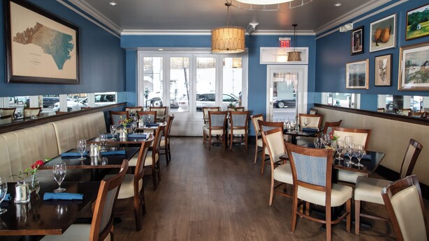 Un restaurant avec des tables et des chaises en bois, un mur bleu orné de tableaux, et une grande fenêtre donnant sur la rue.
