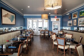 Um restaurante com mesas e cadeiras de madeira, uma parede azul com quadros e uma grande janela com vista para a rua.