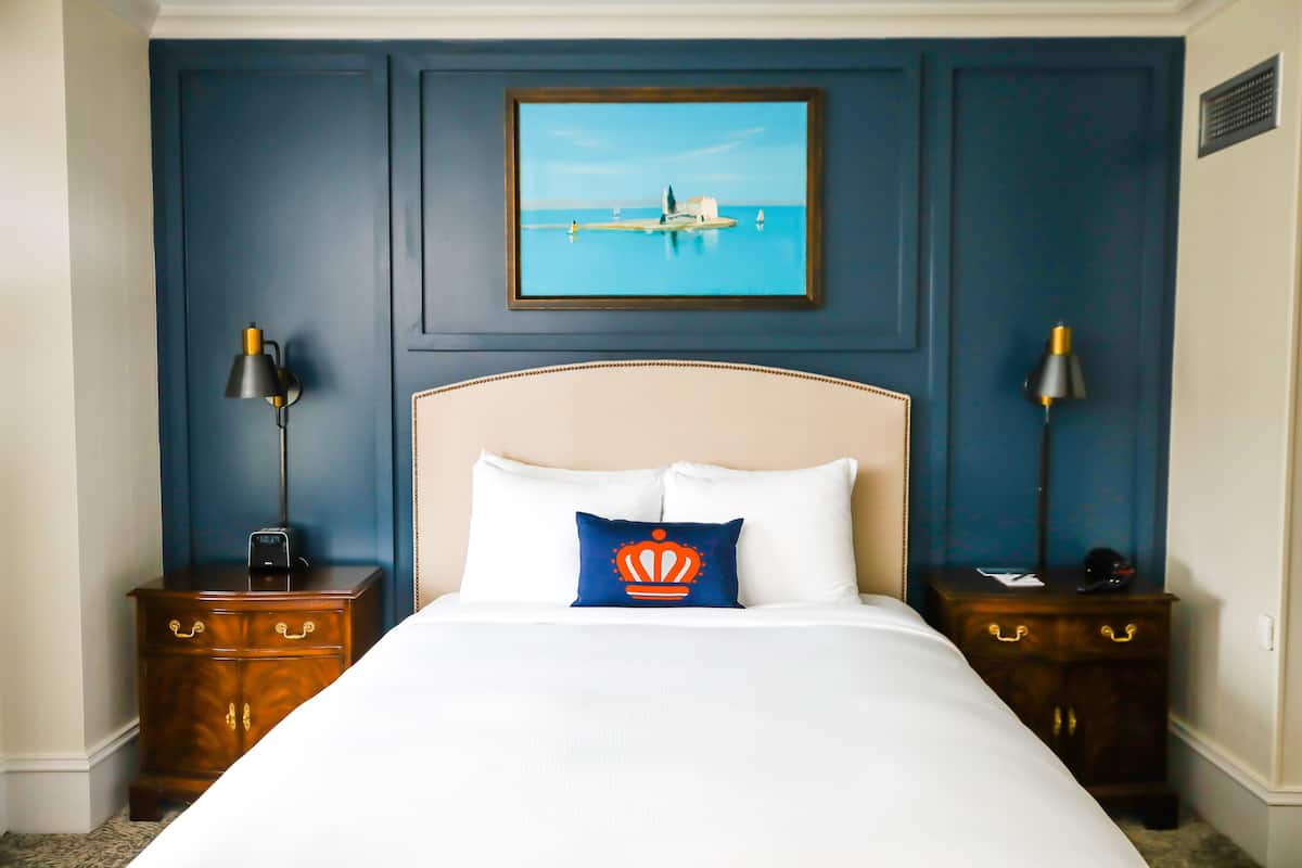 A hotel room with a large bed, two nightstands, a framed painting of a coastal scene, and a decorative pillow.