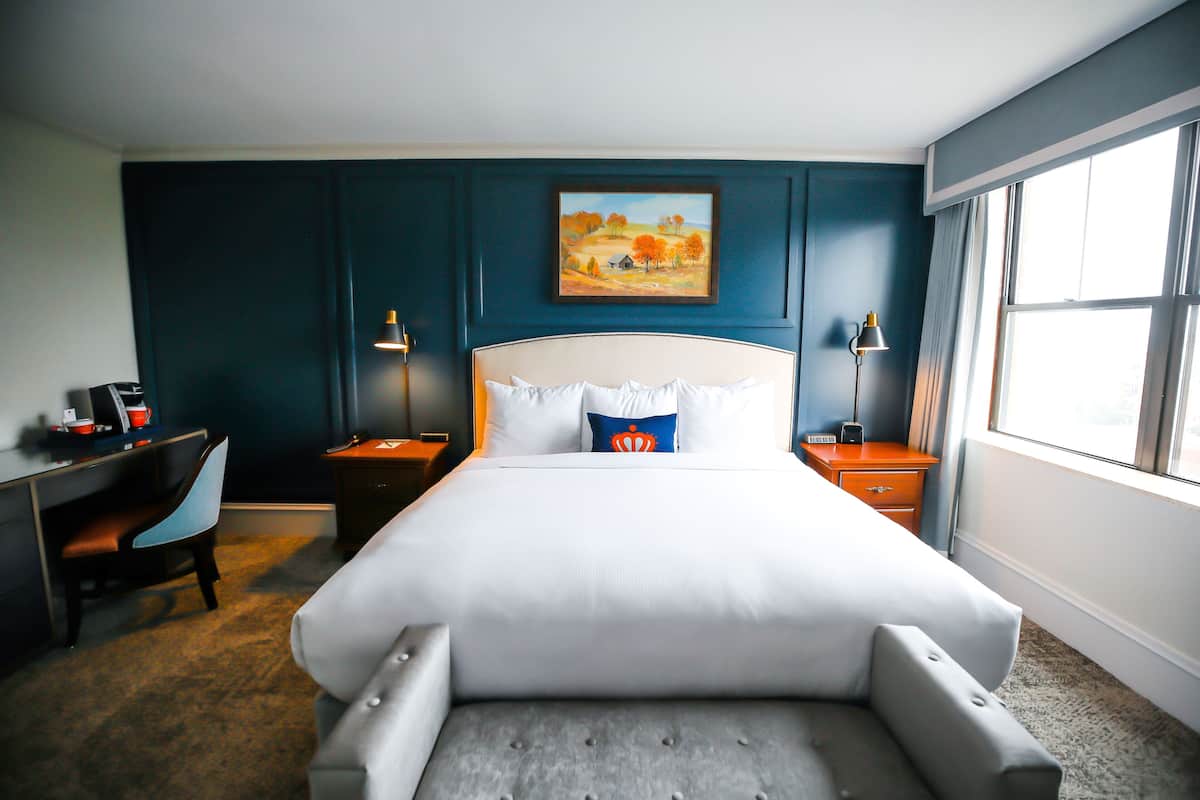 A hotel room with a bed, a desk, a chair, and a painting on the wall.