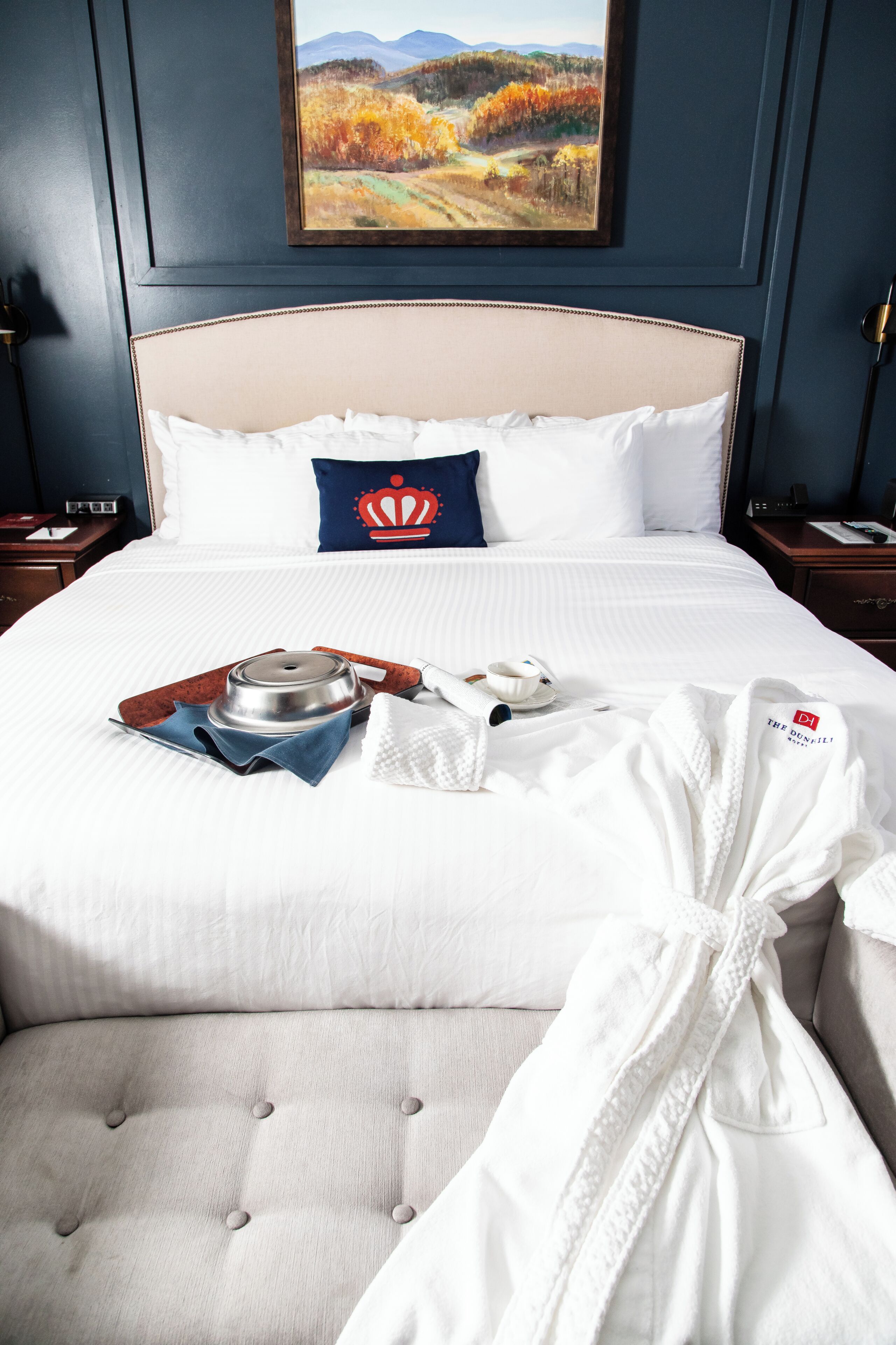 Uma cama bem arrumada com um edredom branco, um travesseiro azul com desenho de coroa e um roupão branco sobre a cama.