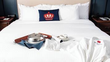 Uma cama bem arrumada com um edredom branco, um travesseiro azul com desenho de coroa e um roupão branco sobre a cama.
