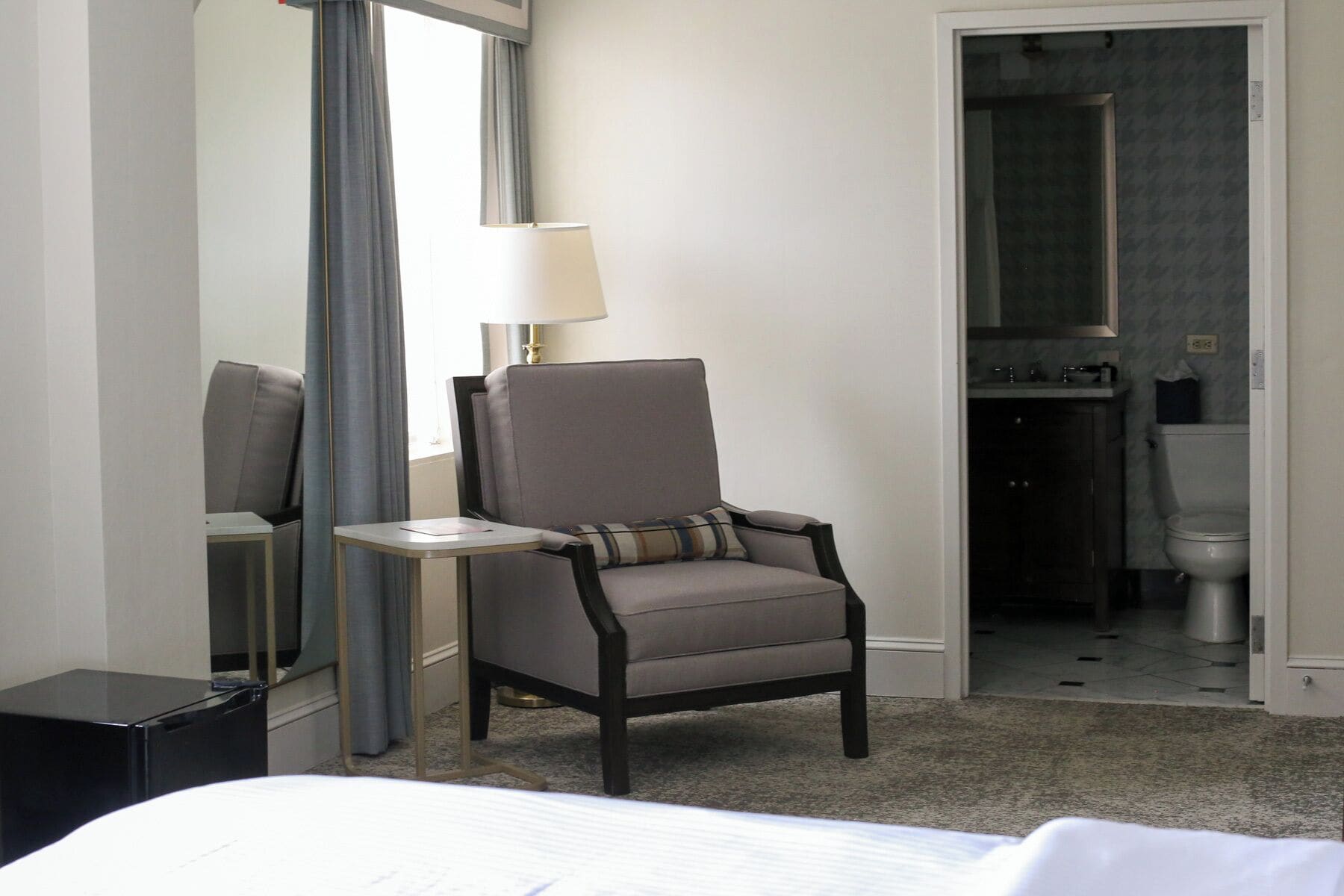 A hotel room with a bed, a chair, a small table, a lamp, and a bathroom visible through an open door.