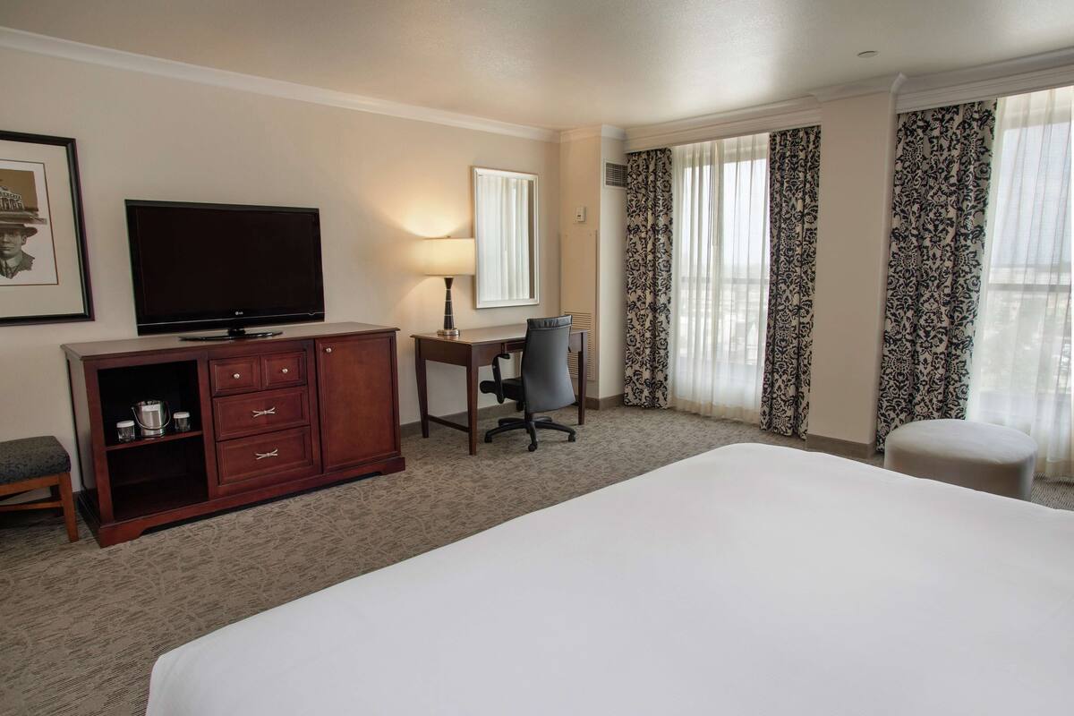 A hotel room with a large bed, a desk with a chair, a television, a dresser, and a window with curtains.