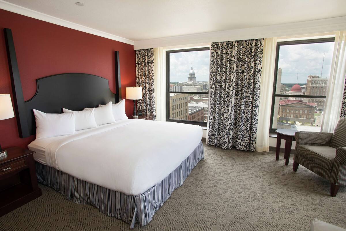 A hotel room with a large bed, a nightstand, a chair, and a view of the cityscape through large windows.