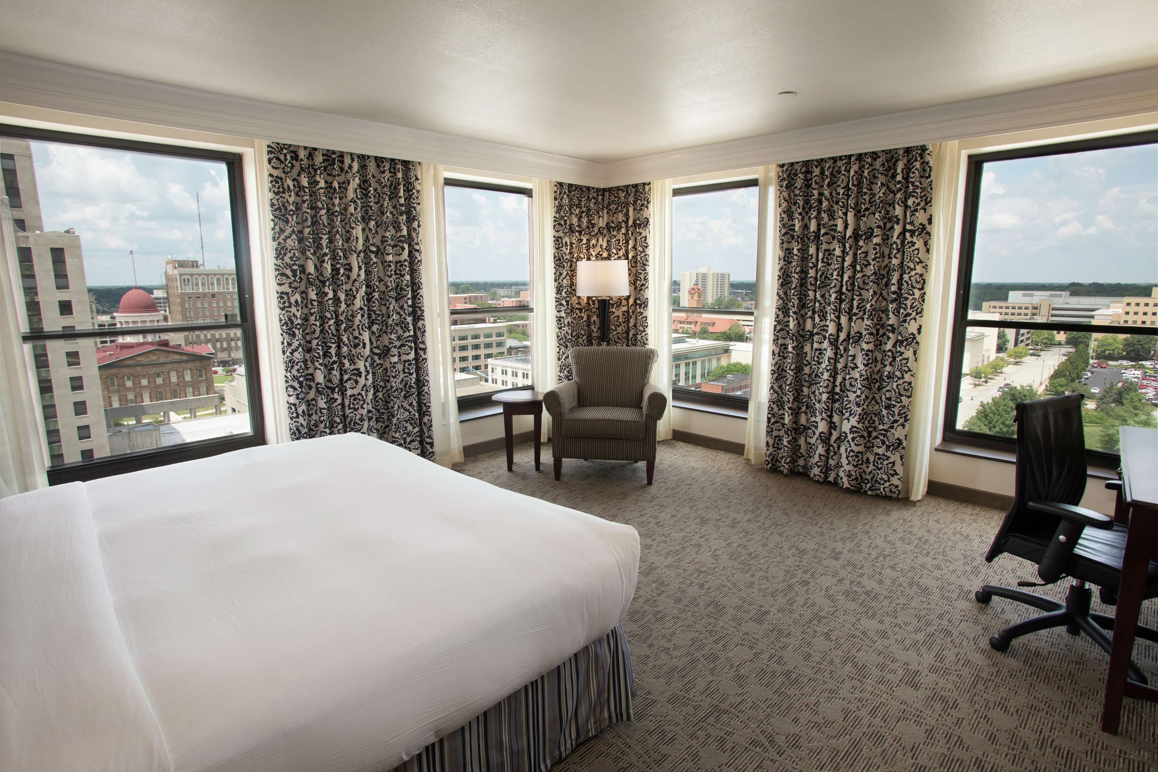 A hotel room with a large bed, a desk, a chair, and a view of the city through large windows.