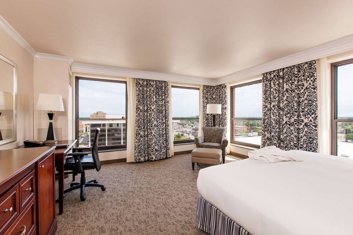 A hotel room with a large bed, a desk with a chair, a small armchair, and a view of the cityscape through large windows.