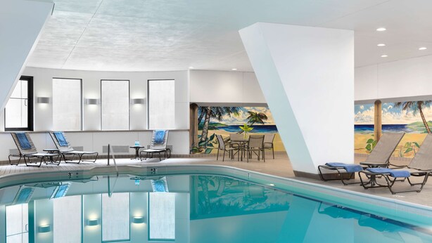 A modern indoor pool area with a clear view of the pool, lounge chairs, and a mural of a beach scene.