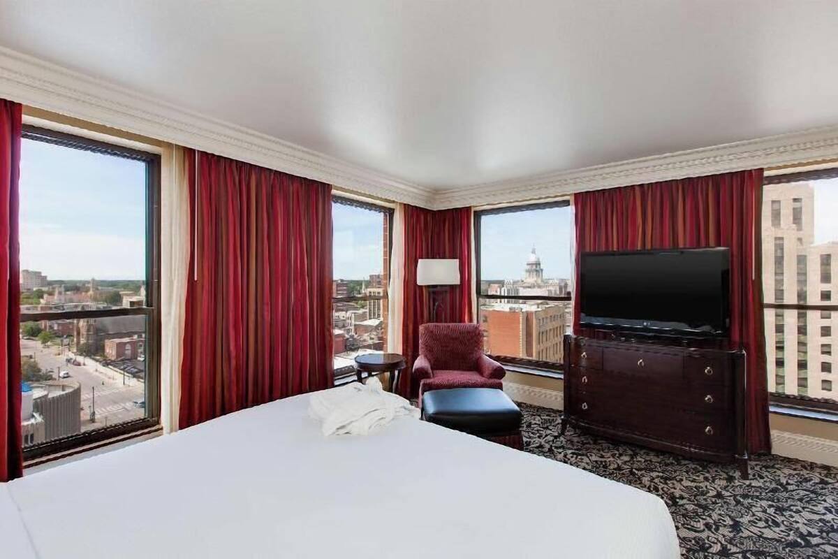 A hotel room with a large bed, a red armchair, a TV, and a city view.