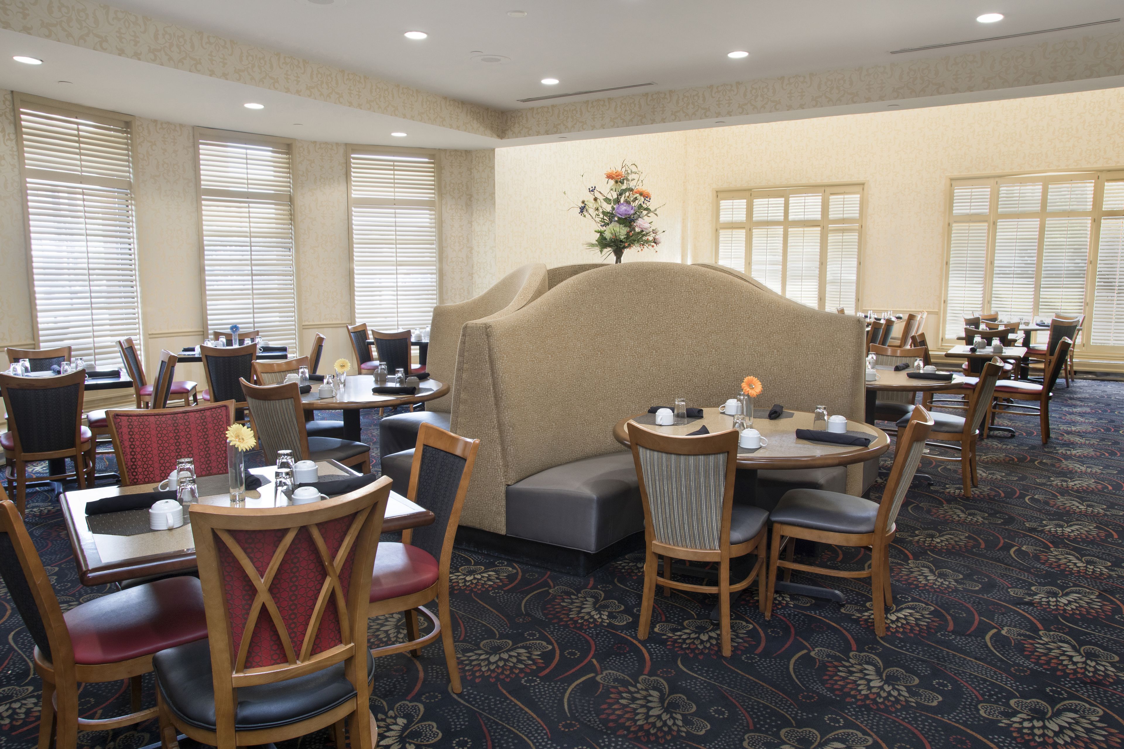 A hotel dining area with multiple tables and chairs, a central seating area, and large windows with blinds.