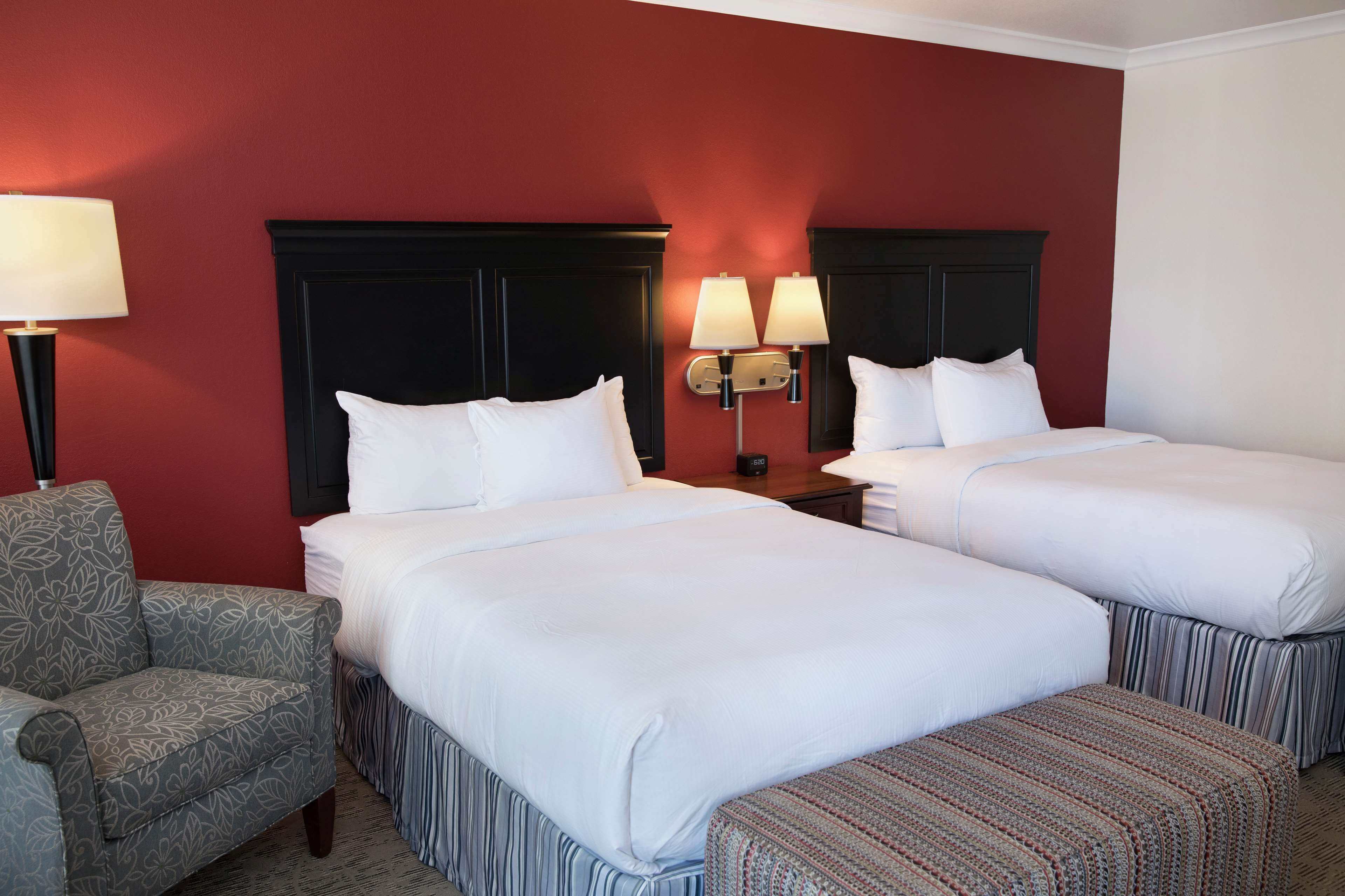 A hotel room with two beds, a nightstand, a lamp, and a patterned armchair.