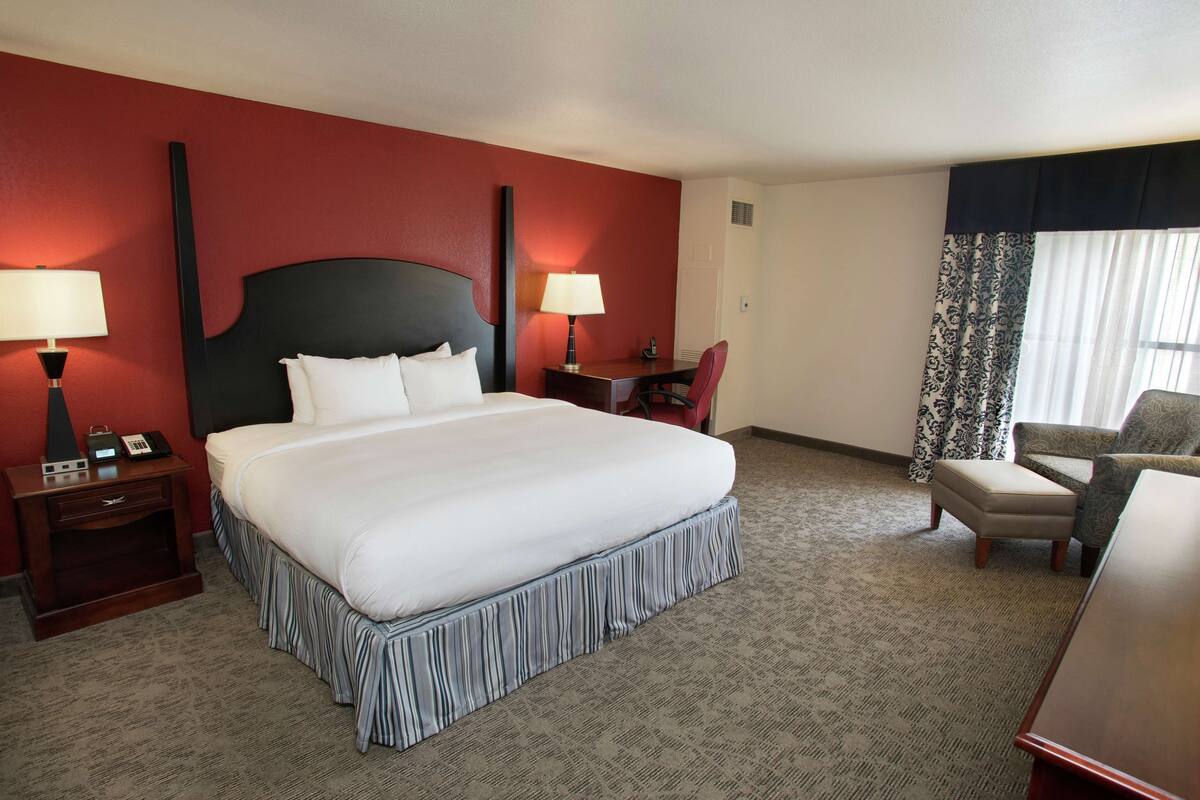 A hotel room with a large bed, two bedside tables with lamps, a desk, a chair, and a window with curtains.