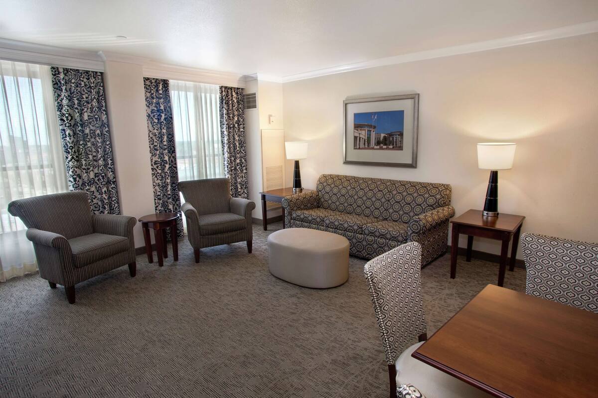 A hotel room with a patterned sofa, two armchairs, a small ottoman, a wooden coffee table, and two bedside tables with lamps.