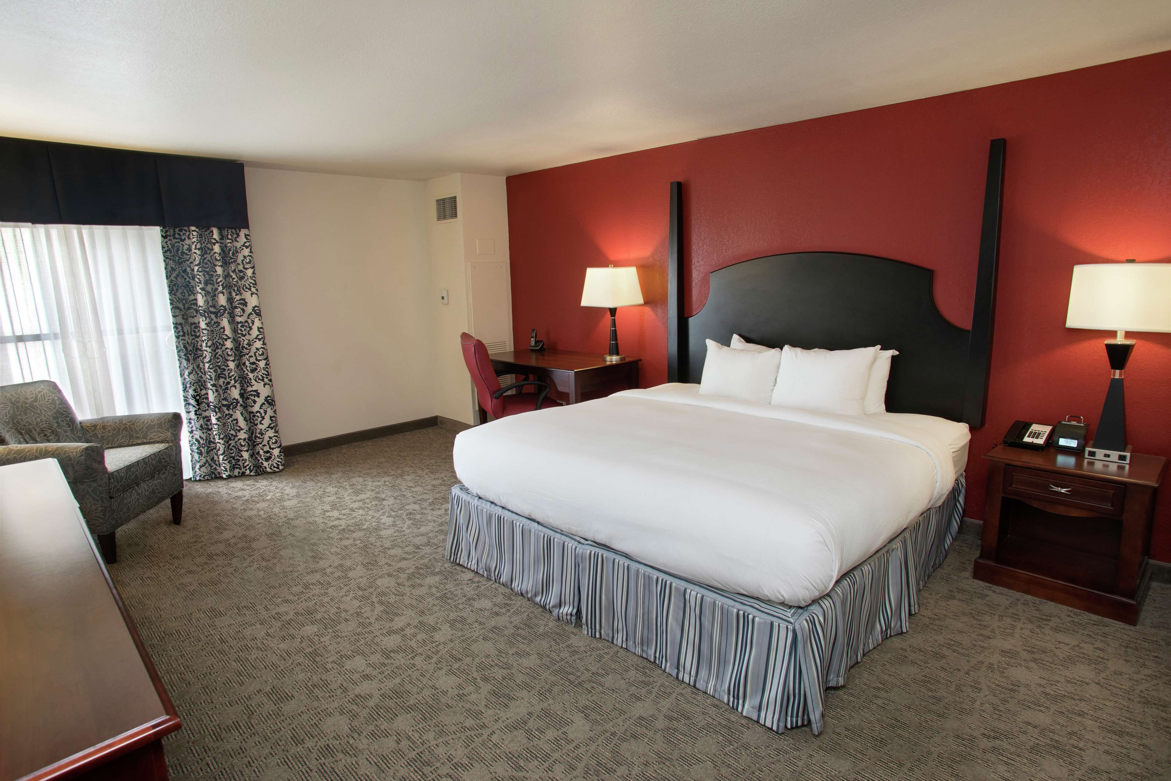 A hotel room with a large bed, two bedside tables with lamps, a desk, a chair, and a patterned curtain.