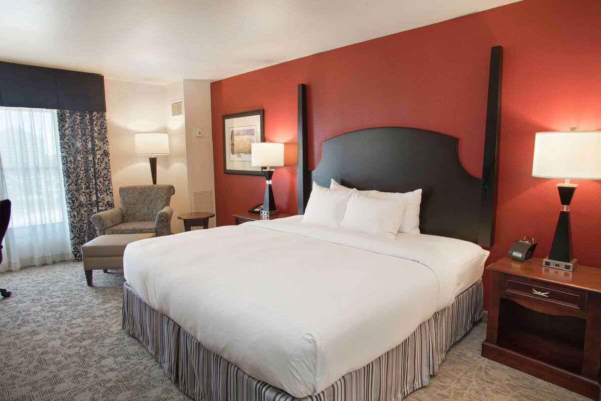 A hotel room with a large bed, a chair, two bedside tables with lamps, a framed picture on the wall, and a window with curtains.