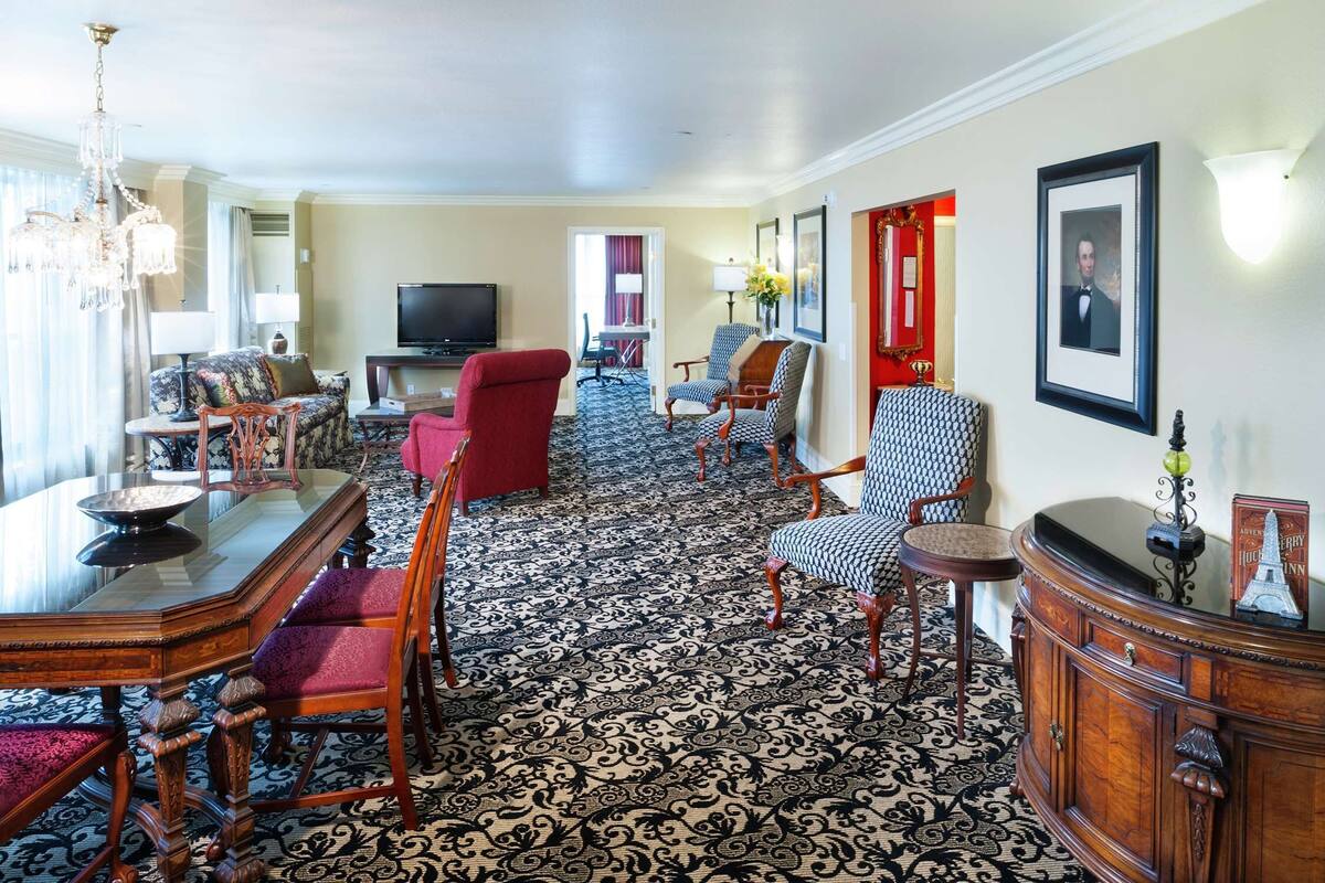 A hotel room with a dining area, a TV, a red armchair, patterned carpet, and a large mirror.