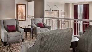 A hotel lobby with a seating area featuring two armchairs and a small table, a decorative wall map, and a view of the lobby through a glass partition.