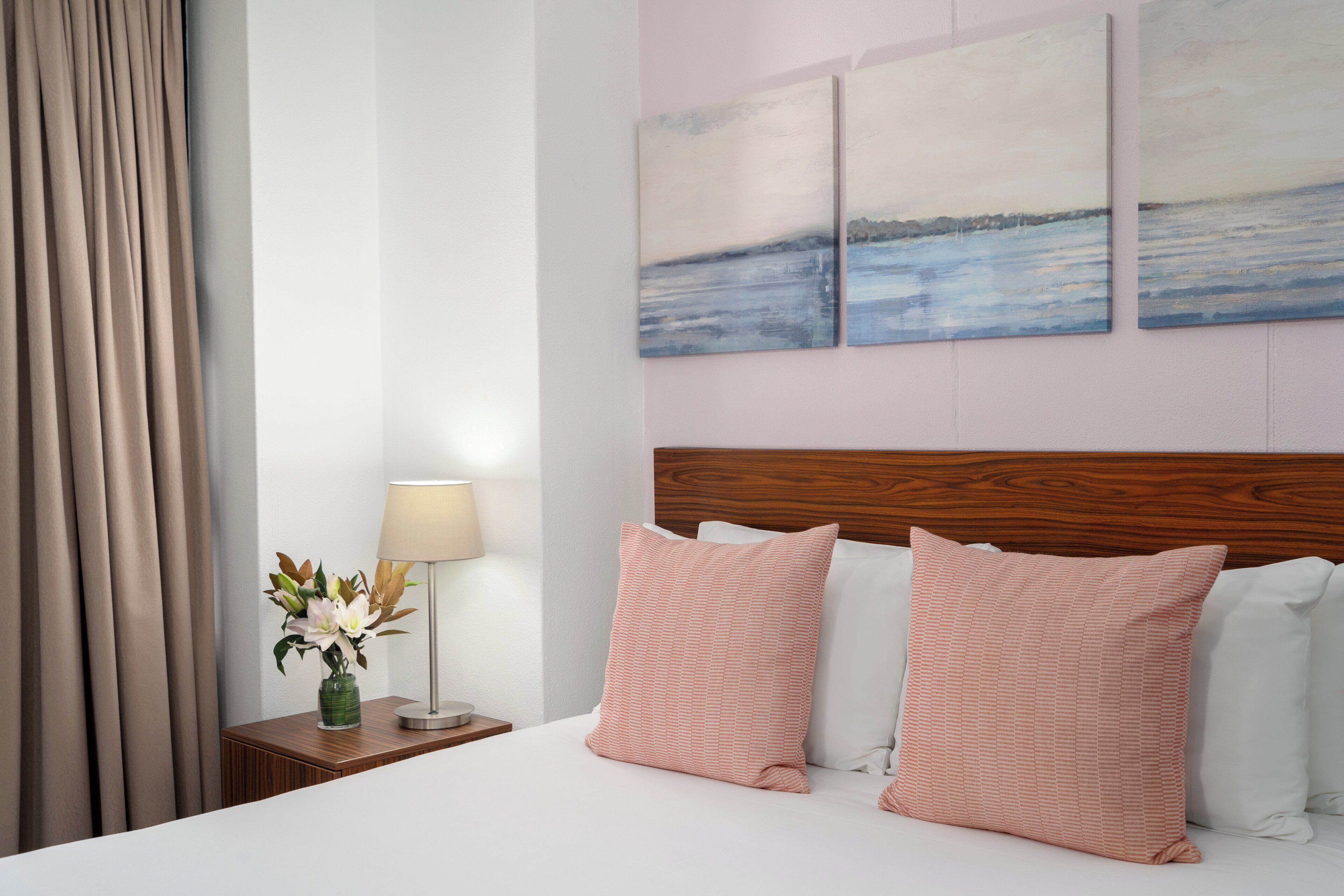 A neatly made bed with two pink pillows, a wooden headboard, a nightstand with a lamp and a vase of flowers, and a view of a lake through the window.
