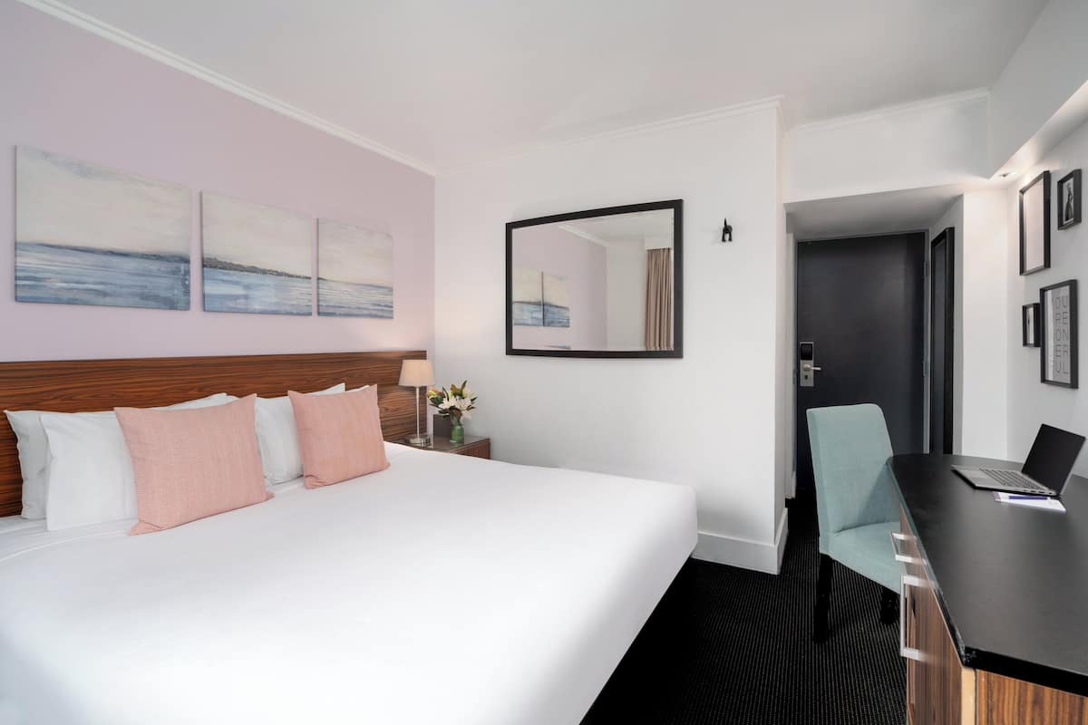 A hotel room with a large bed, a desk with a laptop, a chair, and artwork on the wall.