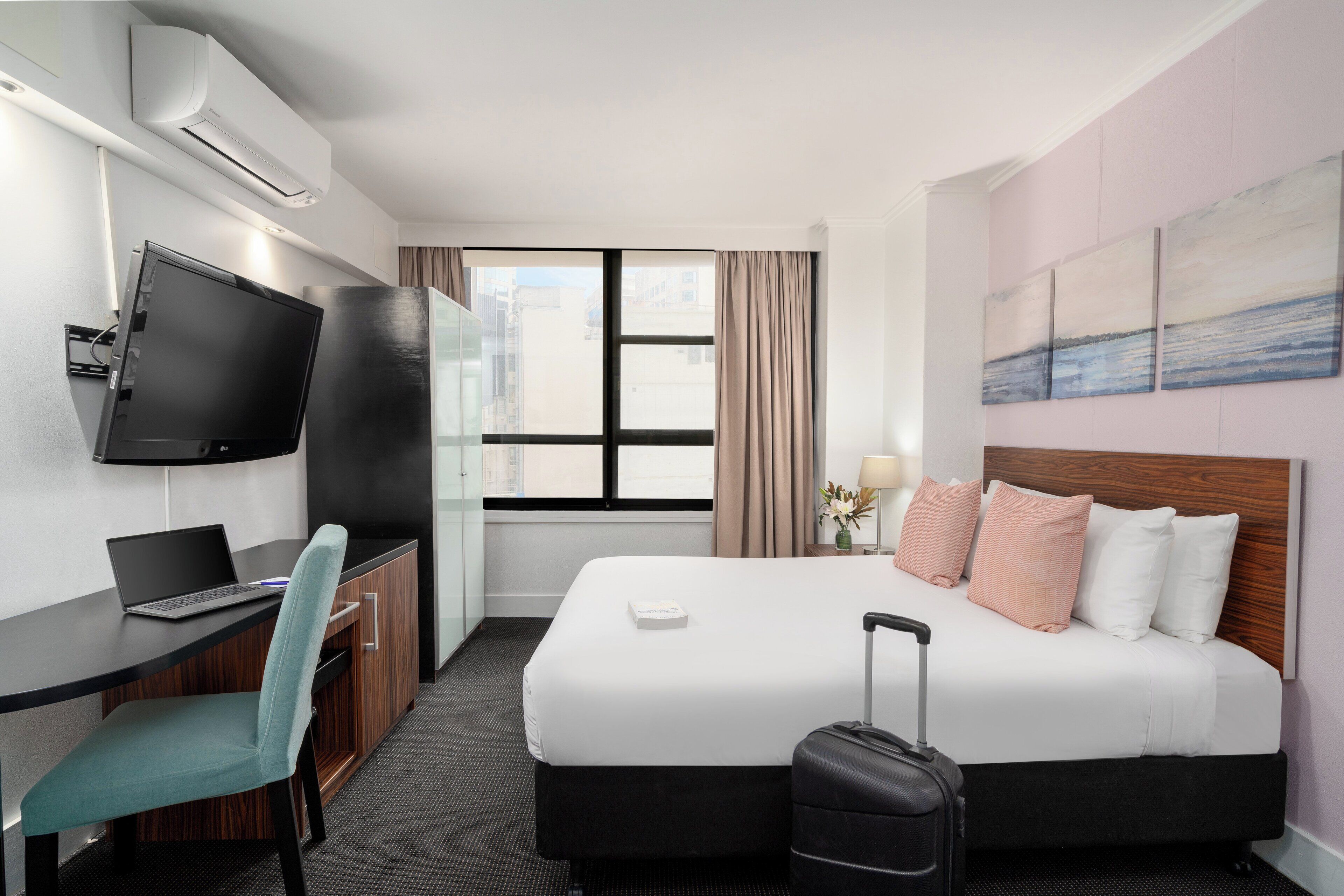 A hotel room with a bed, a desk with a laptop, a television, and a suitcase.
