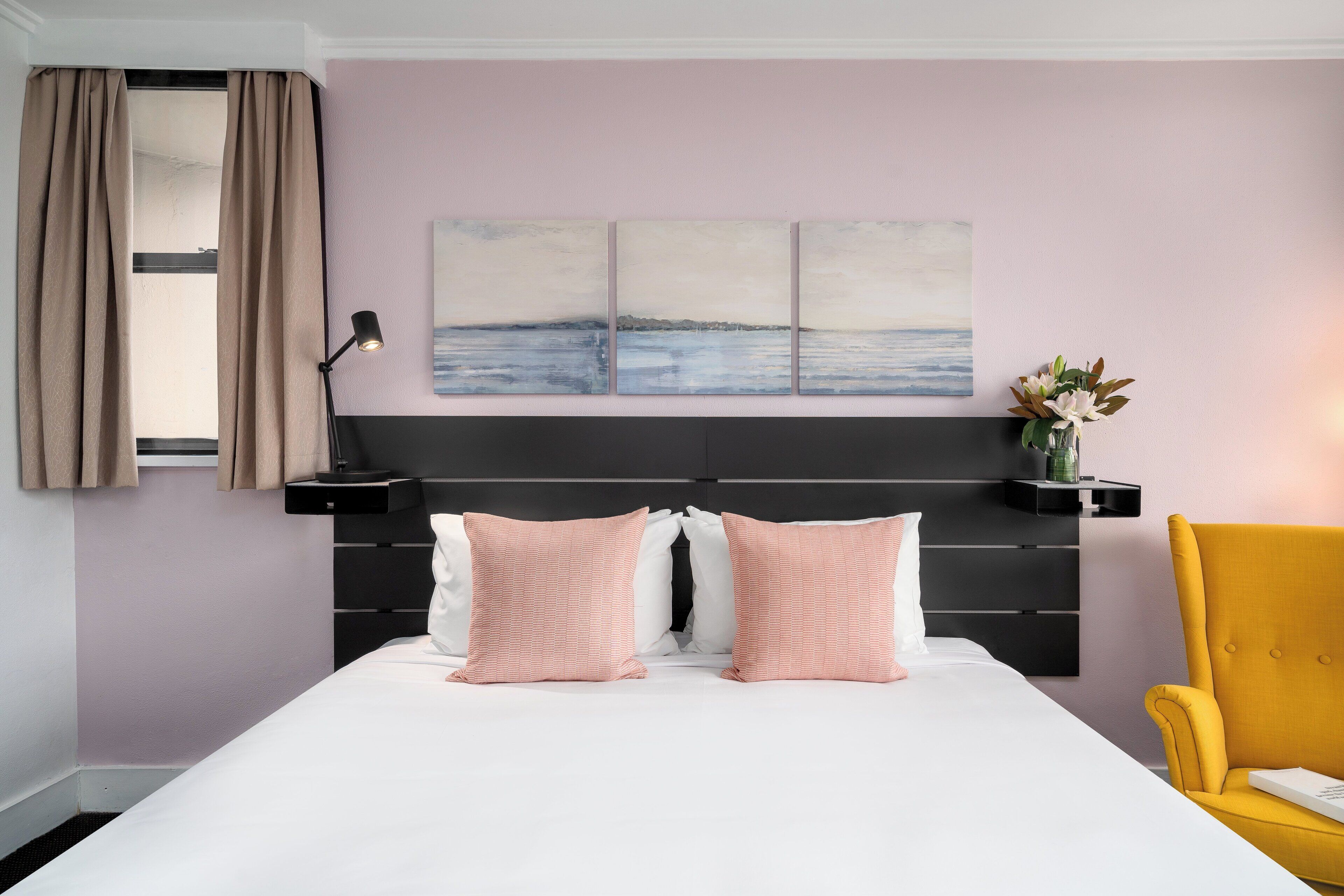 A bedroom with a bed, two pillows, a nightstand, a lamp, a chair, and a wall-mounted shelf with a vase of flowers.