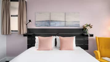 A bedroom with a bed, two pillows, a nightstand, a lamp, a chair, and a wall-mounted shelf with a vase of flowers.