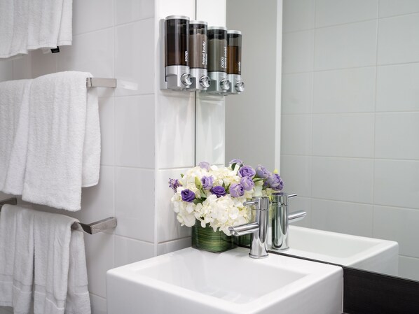 A bathroom with a white sink, a mirror, and a vase of flowers. There are also three soap dispensers on the wall.