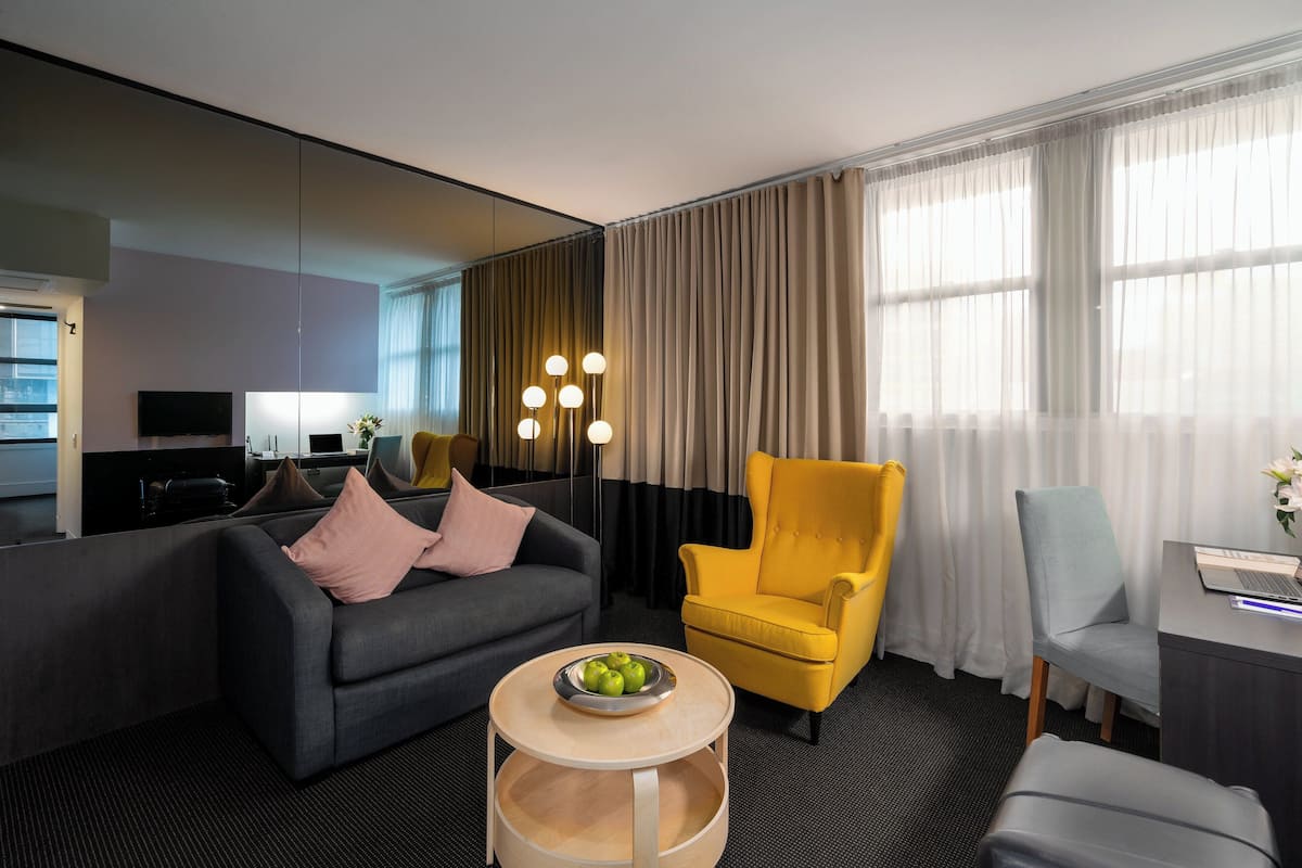 A modern hotel room with a grey sofa, yellow armchair, and a grey armchair. There is a round wooden table with a bowl of fruit. The room has a large mirror, a flat-screen TV, and a desk with a laptop.