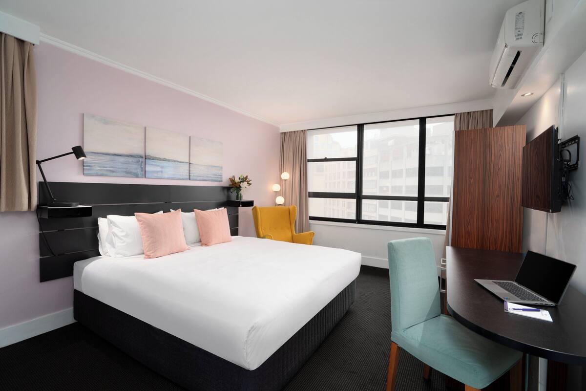 A hotel room with a bed, a desk with a laptop, a chair, a TV, and a window with a city view.